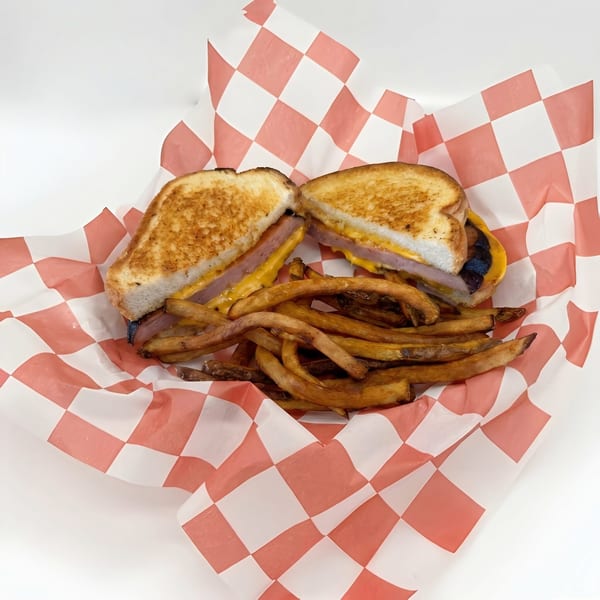 Grilled cheese sandwich with ham and fries in a red and white checkered paper-lined basket.